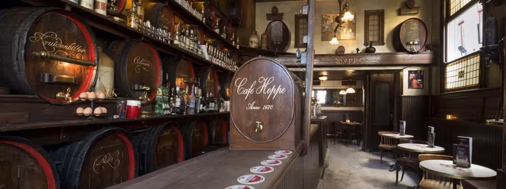 The image showcases the interior of a bar, featuring a prominent barrel with 'Cafe Hoppe' inscribed on it. The composition is decent, with the barrel serving as the focal point. The lighting is adequate, though slightly dim. Sharpness is acceptable, but not exceptional. Color accuracy appears good, with warm tones dominating the scene. The image is reasonably photo-realistic. The key elements, such as the barrel and the bottles behind it, are in focus. The image is not overly cluttered and does not contain any unwanted elements such as watermarks, half-eaten food, or empty drinks. The Amstel coasters on the counter are acceptable. The image is well-composed and lit, with good color accuracy and sharpness. The focus is on the key elements, and the image is photo-realistic. The image is slightly dim, which affects the overall score.