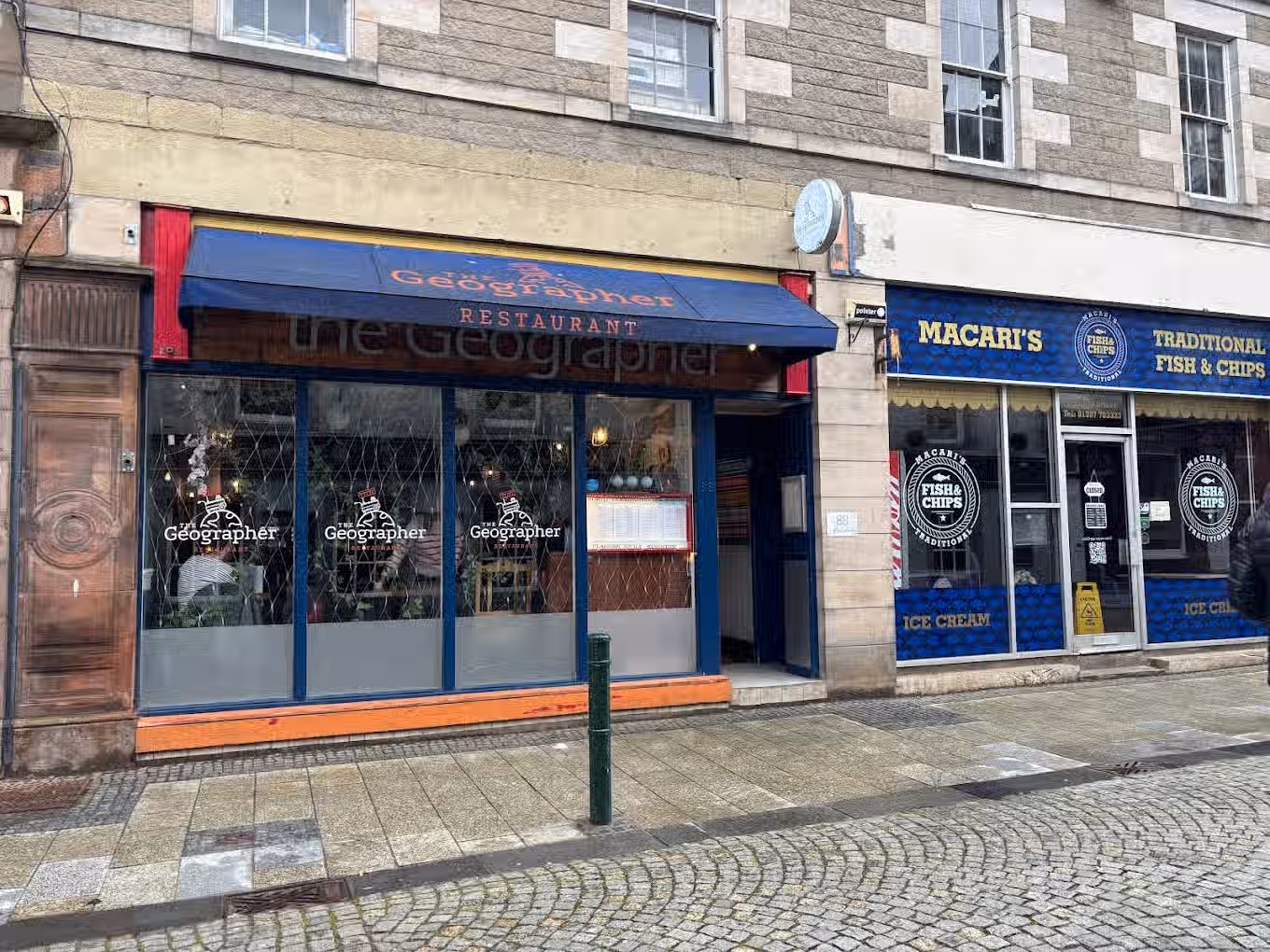 The image shows the exterior of a restaurant called 'The Geographer' and a fish and chips shop called 'Macari's'. The composition is adequate, but not particularly striking. The lighting appears to be natural daylight, which is acceptable. The sharpness is reasonable, but could be improved. The colors seem accurate. The image is photo-realistic. The key elements, such as the restaurant's name and signage, are in focus. The presence of another shop in the frame is not ideal, but acceptable. The image is not particularly well-composed, and the sharpness could be better, but overall it is a decent representation of the scene.