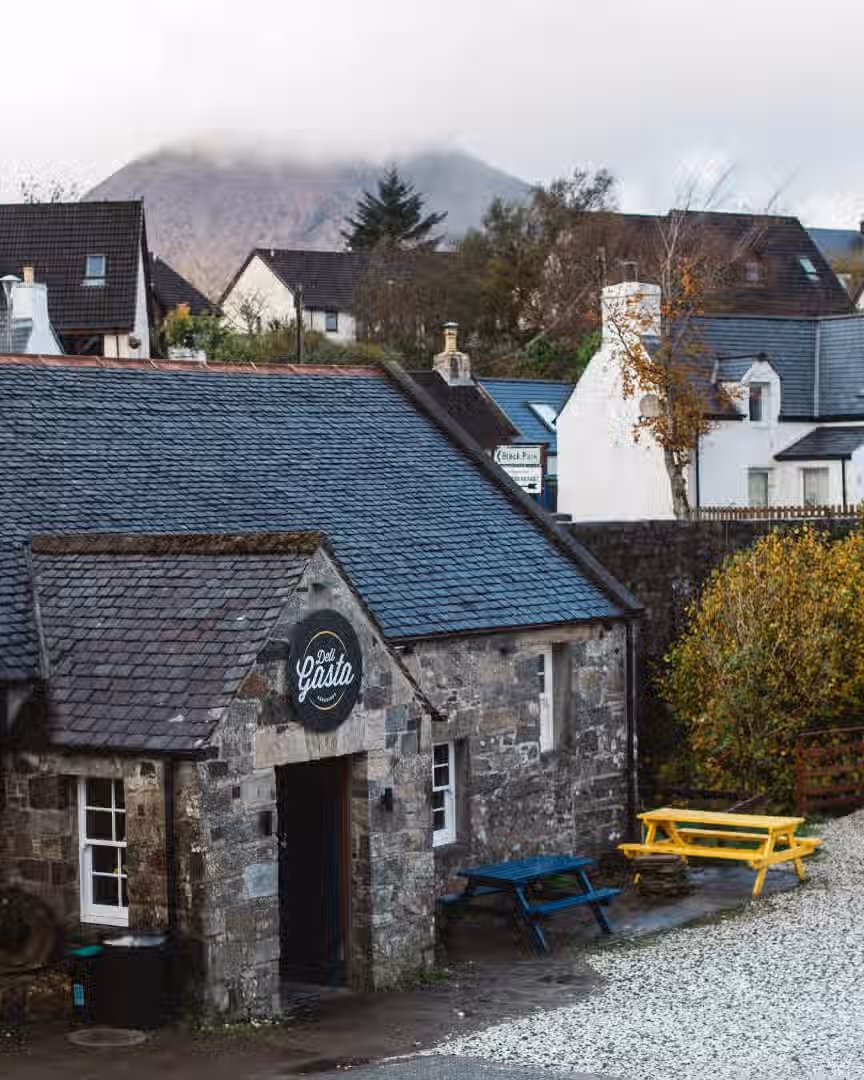 The image showcases a village scene with a stone building in the foreground, a mountain in the background, and colorful outdoor seating. The composition is decent, though slightly tilted. The lighting is adequate, though a bit flat due to overcast conditions. Sharpness is acceptable, but not exceptional. Color accuracy seems fine. The image is reasonably photo-realistic. The key elements are mostly in focus. The overall quality is good, but improvements could be made in composition and sharpness.