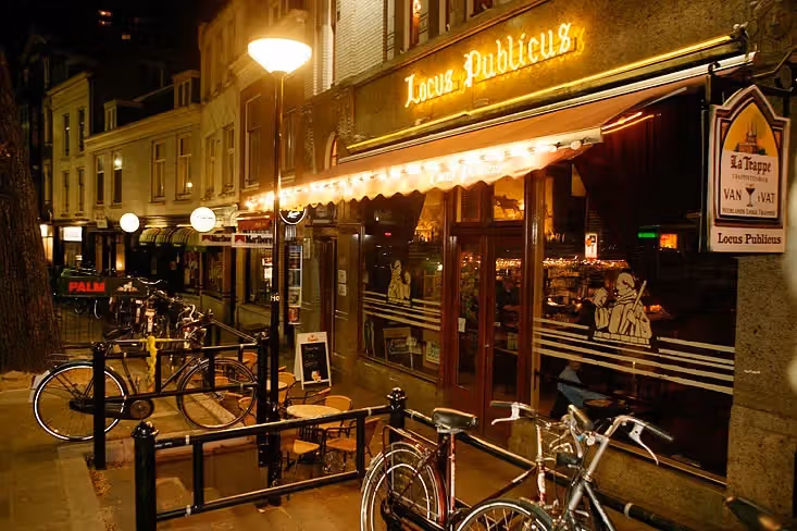 The image shows the exterior of a bar or restaurant at night. The composition is decent, but the presence of bicycles in the foreground is somewhat distracting. The lighting is adequate, with the neon sign and street lamps providing illumination. The image is not very sharp, and the color accuracy is acceptable, though the overall tone is warm. The image is somewhat photo-realistic. Key elements are reasonably in focus. The presence of a Marlboro advertisement is an allowed element. The overall image quality is acceptable, but could be improved with better composition and sharpness.