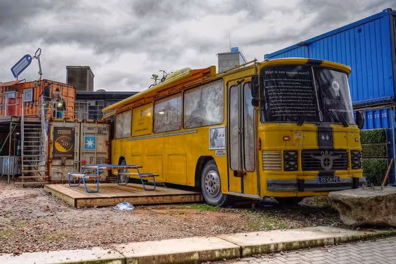 The image shows a yellow bus converted into an escape room, parked next to shipping containers. The composition is decent, but the overall sharpness is lacking, and the image appears slightly processed, impacting the photo-realism. The lighting is adequate, though the cloudy sky results in somewhat flat lighting. Color accuracy seems acceptable. The key elements, like the bus and the text on the window, are reasonably in focus. The image is well-composed. However, it is not a professional photograph and has some distortion. The image has a unique and creative element, but the overall image quality is not the best.