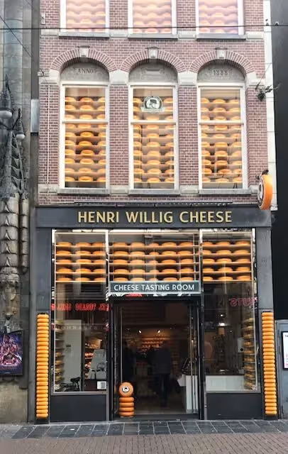 The image shows the storefront of a cheese shop called Henri Willig Cheese. The composition is decent, focusing on the building's facade. The lighting appears to be natural daylight, which is adequate but not exceptional. The sharpness is acceptable, though finer details could be crisper. The colors seem accurate, with the cheese wheels appearing a natural yellow-orange. The image is reasonably photo-realistic. Key elements, like the sign and the cheese wheels, are in focus. It is well-composed and the image is not cluttered. The image quality is good, but could be improved with better lighting and sharpness. The image serves as an advertisement for the cheese shop.