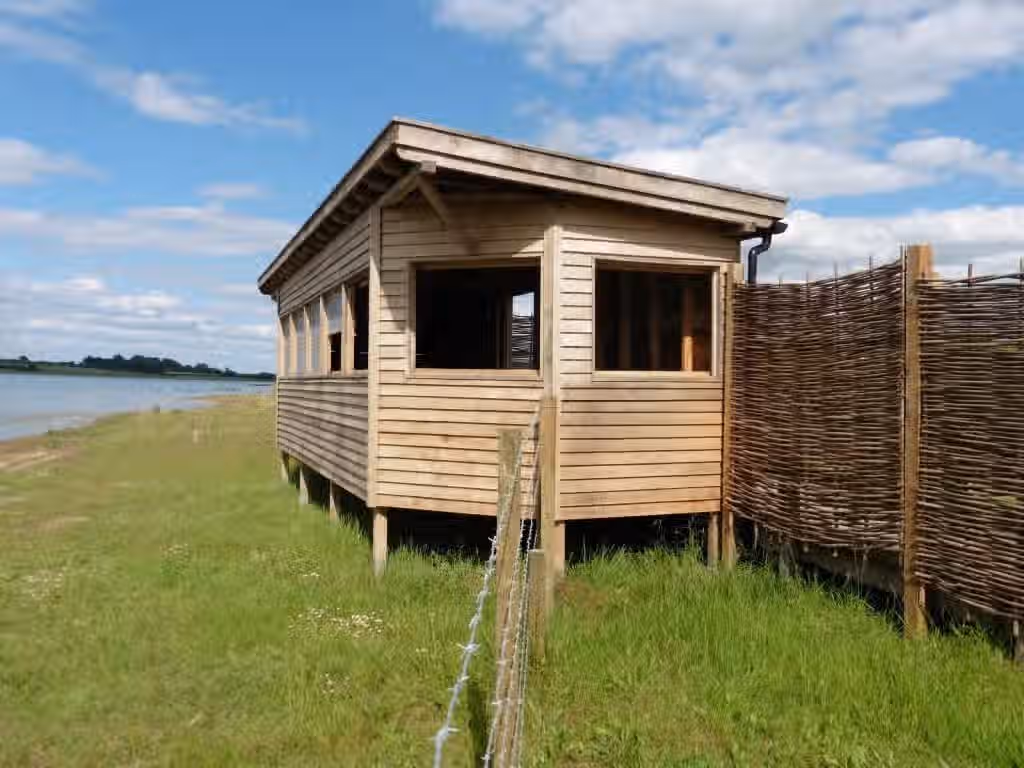 The image shows a wooden structure in a grassy field. The composition is decent, but could be improved. The lighting is good, and the colors appear accurate. The sharpness is acceptable, but not exceptional. The image appears photo-realistic. The key elements, such as the building, are generally in focus. The barbed wire fence in the foreground is a slightly unwanted element, but not enough to bring the score to 0. Well-lit and color accurate, but the composition and sharpness could be improved.