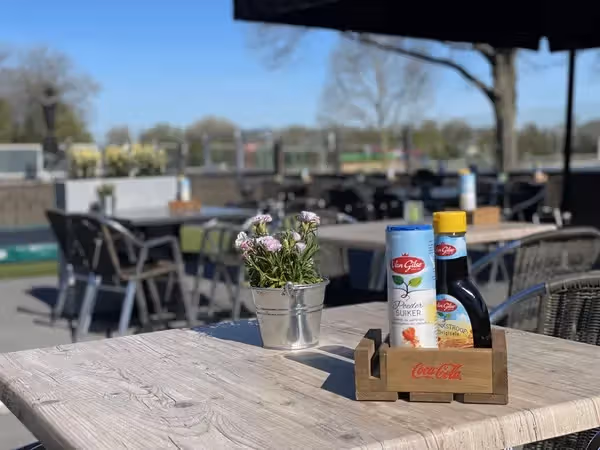 The image showcases a table setting at an outdoor cafe or restaurant. The composition is decent, with the main elements (flowers, sugar, syrup) in focus. The lighting is good, but the image could be sharper. The colors are accurate, and the image appears photo-realistic. Key elements are in focus. The presence of promotional material (Coca-Cola logo) is allowed. The background is slightly blurred, which helps to draw attention to the foreground elements. Overall, a presentable image, but with room for improvement in sharpness and composition.