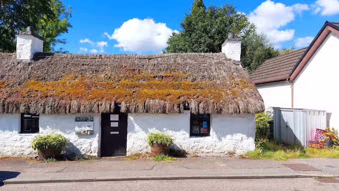 The image shows a charming cottage with a thatched roof. The composition is decent, although slightly off-center. The lighting is good, with bright daylight and clear skies. The sharpness is acceptable, but could be improved. The colors appear accurate. The image is reasonably photo-realistic. The key elements, such as the cottage and roof, are in focus. Overall, the image is well-executed and visually appealing. The presence of signage and the plant pot are acceptable elements. The composition could be improved to better highlight the cottage. The image is well-lit and color accurate. Sharpness is good, but could be better to capture finer details of the thatched roof. The image is photo-realistic.