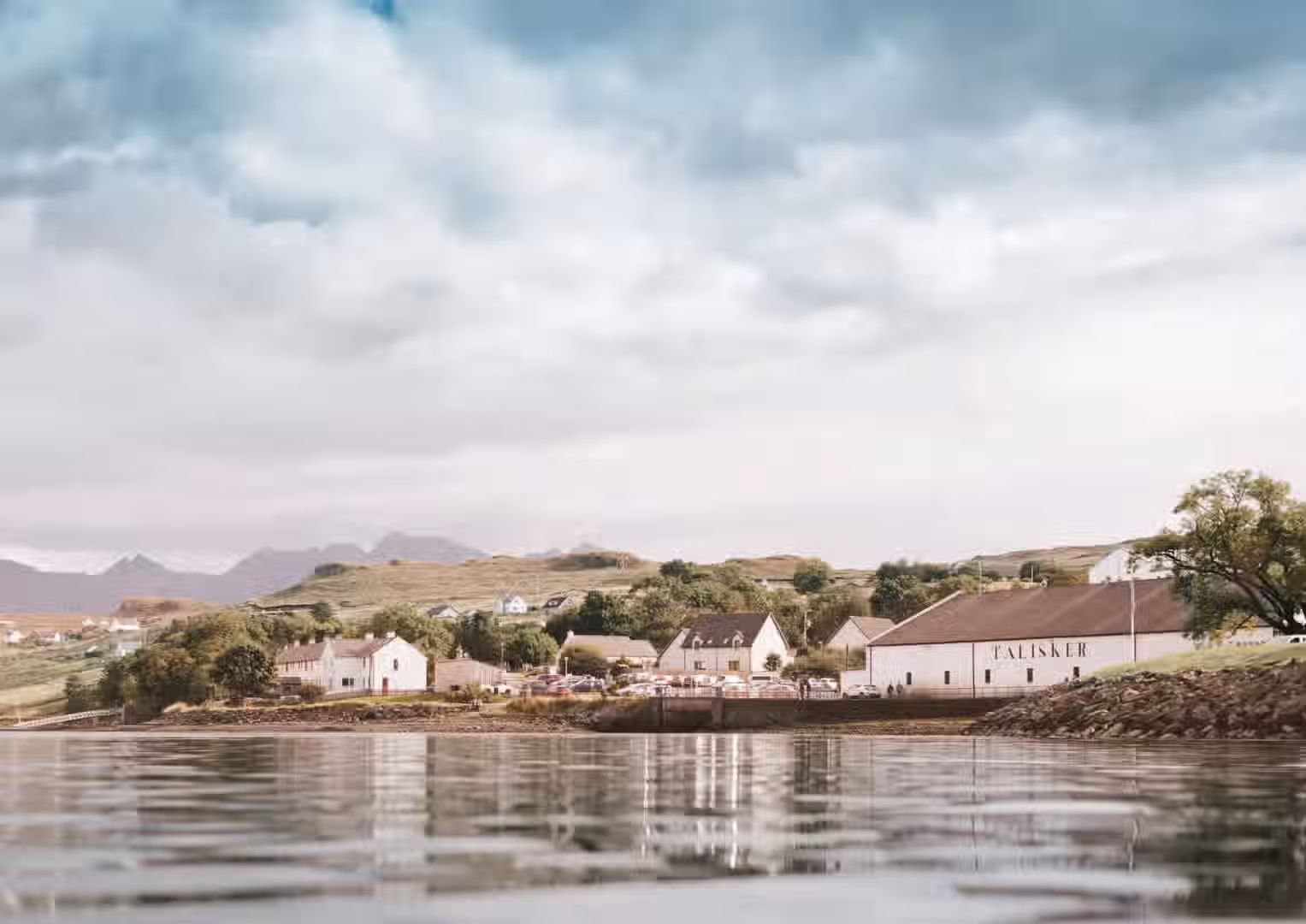 The image depicts a coastal town with buildings reflected in the water. The composition is decent, with the buildings and landscape elements arranged in a visually appealing manner. The lighting is soft, likely due to overcast conditions, which contributes to a subdued but pleasant atmosphere. The sharpness is acceptable, though some finer details could be more crisp. The colors appear accurate. The key elements, such as the buildings and their reflections, are in focus. The presence of the Talisker distillery adds a unique element to the scene. Overall, the image is well-composed and visually appealing, with minor deductions for sharpness and slightly muted colors.
