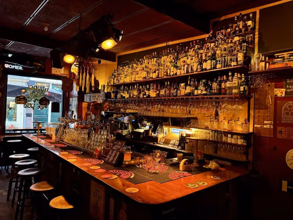 The image showcases the interior of a bar, featuring a well-stocked liquor shelf, glasses, and a counter. The composition is decent, capturing a good portion of the bar's ambiance. The lighting is somewhat dim, contributing to a warm, inviting atmosphere, but it could be brighter for better visibility. The sharpness is acceptable, but could be improved. The color accuracy seems reasonable. The image is reasonably photo-realistic. Key elements like the bottles and glasses are mostly in focus. Overall, the image is well-composed and conveys the atmosphere of the bar effectively, but could benefit from improved lighting and sharpness. It doesn't contain any unwanted elements.