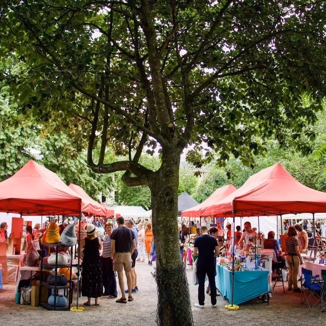 The image depicts an outdoor market scene with red tents and people browsing. The composition is somewhat obstructed by the tree in the foreground. The lighting is adequate but slightly uneven due to the tree's shade. Sharpness is acceptable but not exceptional. Color accuracy appears reasonable. The photo-realism is good. Key elements, such as the people and tents, are generally in focus. The composition could be improved by positioning the tree off to the side. The image is not overly cluttered, but the tree does detract from the overall visual appeal. Well-composed (18/25), Well-lit (12/15), Sharp (14/20), Color-accurate (8/10), Photo-realistic (16/20), Key elements in focus (0/10).