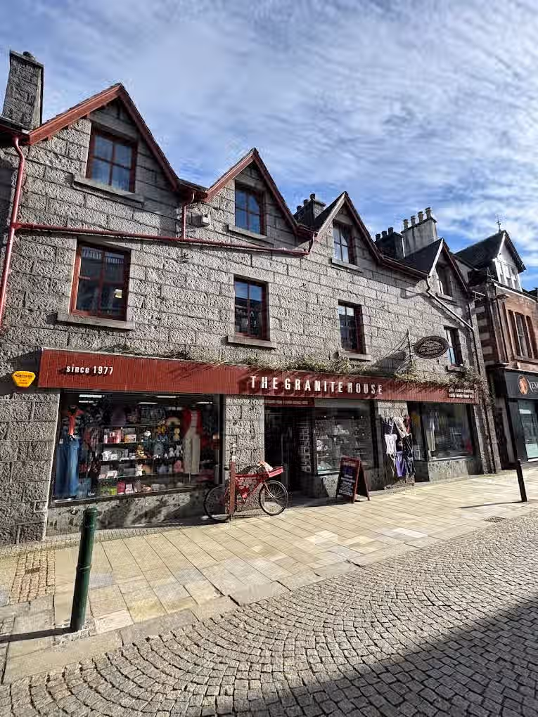 The image shows the exterior of a shop called 'The Granite House'. The composition is decent, capturing the building and its surroundings. The lighting is good, with natural light illuminating the scene. The image is reasonably sharp, although it could be sharper. The colors appear accurate. The image is photo-realistic. The key elements, such as the shop front and the building's facade, are in focus. The presence of a bicycle parked in front of the shop is slightly distracting but not detrimental. Overall, the image is well-presented and informative.