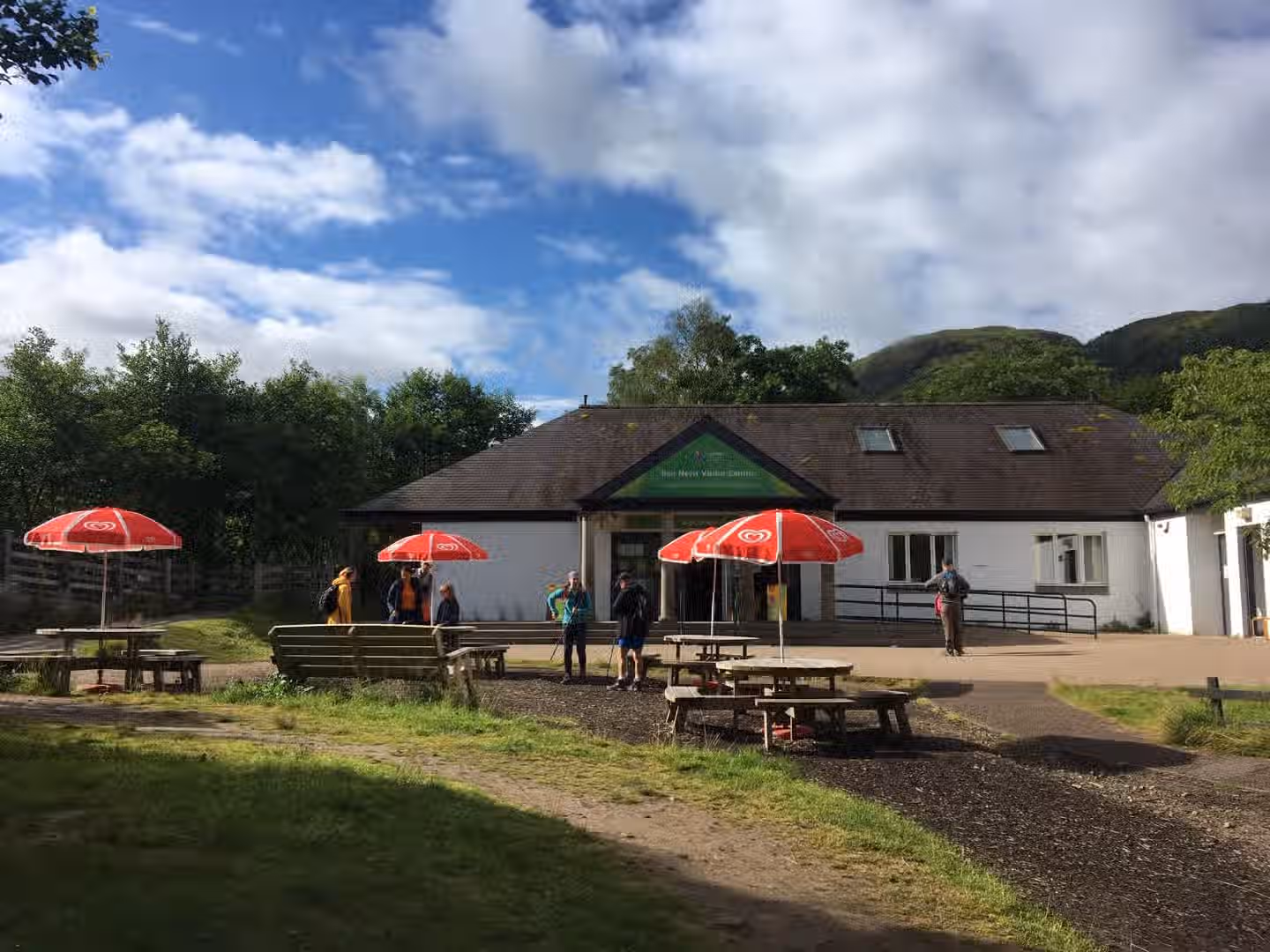 The image shows the exterior of a visitor centre. The composition is acceptable but not perfect. The lighting is adequate, though slightly uneven, with some areas appearing brighter than others. The sharpness is decent, but not exceptional. The colors appear mostly accurate. It doesn't appear particularly photo-realistic, and the key elements, such as the building and the people, are generally in focus. The presence of people adds to the scene but also introduces some potential distractions. The overall impression is that it is a snapshot rather than a professionally composed photograph. The image could benefit from better composition and more even lighting. The image is not well-composed and the sharpness is not great. The image is not particularly photo-realistic.