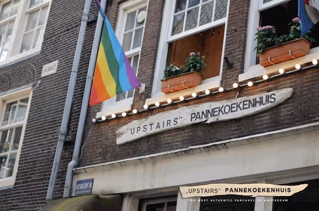 The image shows the exterior of a building, likely a restaurant or pancake house, with a rainbow flag displayed. The composition is acceptable, but the image is not particularly sharp. The lighting is decent, but the colors appear slightly muted. The focus is reasonably good on the central elements. The image is not particularly photo-realistic, and the presence of text and a logo at the bottom detracts from the overall quality. The image is well-composed, but the sharpness and color accuracy could be improved. The key elements are in focus, but the overall image quality is not exceptional. The image is well-lit but not perfectly so. The image is not particularly photo-realistic.