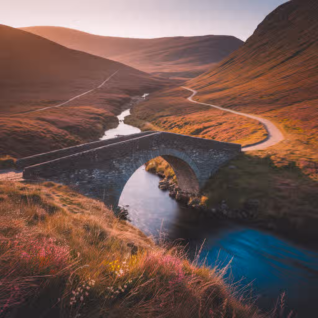 The image is well-composed, showcasing a stone bridge over a river in a mountainous landscape. The lighting is warm and appealing, likely captured during sunrise or sunset. The sharpness is good, with the bridge and surrounding landscape details clearly visible. The colors are accurate and vibrant, enhancing the overall aesthetic appeal. The image appears photo-realistic, with a natural and authentic feel. Key elements, such as the bridge and river, are in focus, drawing the viewer's attention. The composition makes good use of leading lines with the road and river. Overall, the image is of high quality and visually pleasing.