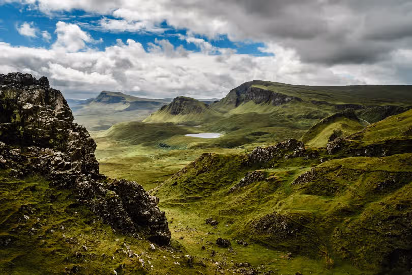 The image presents a well-composed landscape featuring green hills and mountains under a cloudy sky. The lighting is generally good, though slightly muted due to the cloud cover. The sharpness is acceptable, allowing for decent detail in the landscape. The colors appear accurate, with vibrant greens. The image is photo-realistic and the key elements, such as the hills and mountains, are in focus. The composition is strong, guiding the eye through the scene. Overall, the image is visually appealing and of good quality.