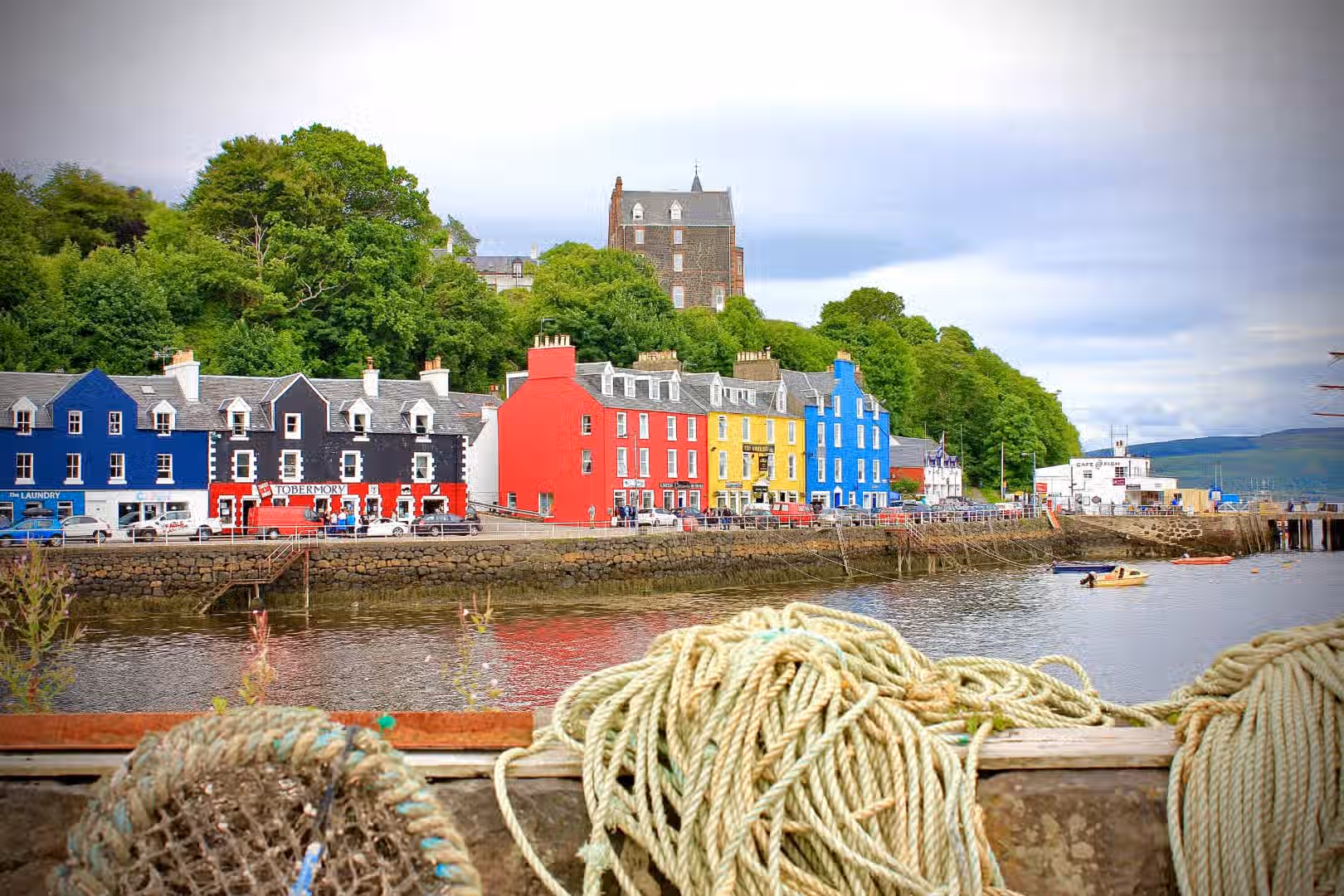 The image depicts the colorful buildings of Tobermory, Scotland, along the waterfront. The lighting is adequate, and the colors are vibrant. The sharpness could be improved, and the focus is slightly distracted by the rope in the foreground. The composition includes a castle in the background and a harbor scene, adding to the context. The image is reasonably photo-realistic. The rope in the foreground is a bit distracting and doesn't add much to the overall image. The key elements (buildings and castle) are somewhat in focus, but not perfectly sharp.