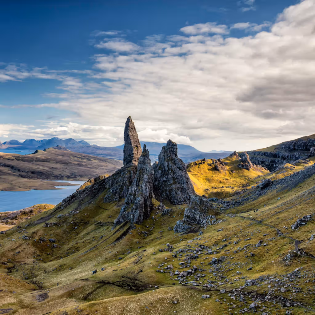 The image showcases a scenic landscape featuring mountains, a lake, and the Old Man of Storr in the Isle of Skye, Scotland. The composition is well-balanced, with the rock formations serving as a strong focal point. The lighting is generally good, with a mix of sunlight and clouds creating interesting shadows and highlights. The image is sharp and detailed, capturing the textures of the rocks and vegetation. The colors appear accurate and vibrant. The key elements, particularly the Old Man of Storr, are in focus. The image is photo-realistic and visually appealing. Strengths: Well-composed, well-lit, sharp, color-accurate, photo-realistic, key elements in focus. Weaknesses: None apparent.