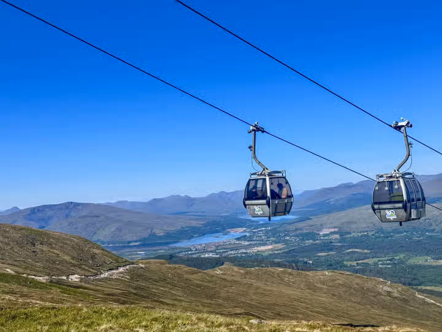 The image is well-composed, capturing a cable car against a scenic mountain landscape. The lighting is bright and even, contributing to a clear and vibrant picture. The image is reasonably sharp, with the cable car and surrounding landscape details visible. Colors appear accurate, with a natural representation of the sky, mountains, and vegetation. The image is photo-realistic and the key elements, particularly the cable car, are in focus. Strengths: well-lit, color-accurate, and the key elements are in focus. Weaknesses: could be sharper.