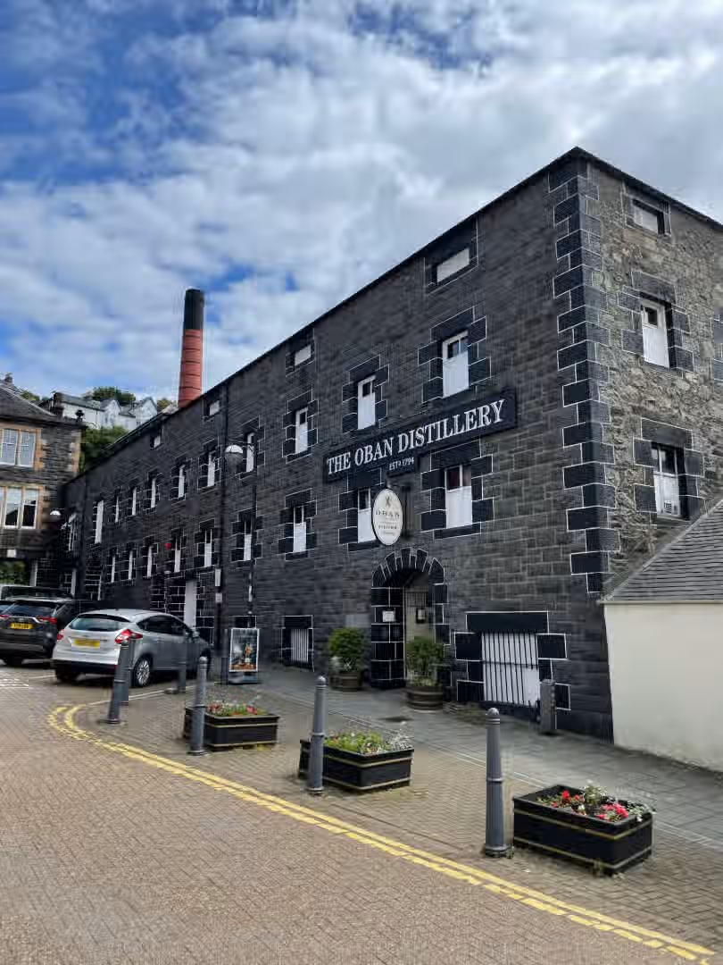 The image shows the exterior of the Oban Distillery. The composition is decent, although the cars in the foreground are somewhat distracting. The lighting is adequate, and the colors appear accurate. Sharpness is acceptable but not exceptional. The key elements, such as the distillery building and signage, are in focus. The image is fairly photo-realistic. The presence of cars could be considered an unwanted element, but they are not significantly detrimental. Overall, the image is acceptable but could be improved with better composition and sharpness.