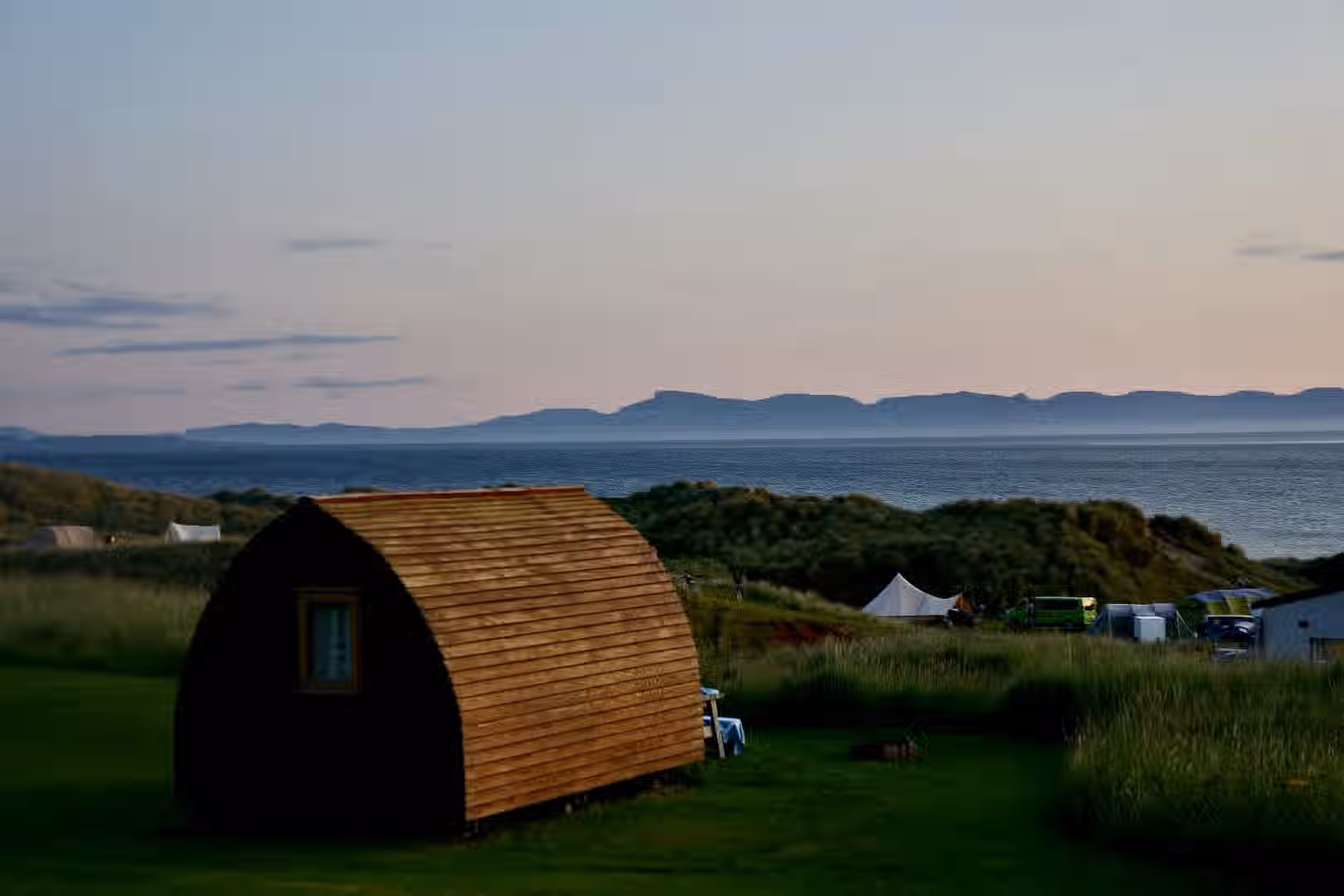 The image shows a cabin in a landscape setting with the ocean and mountains in the background. There is also a tent visible. The composition is decent, but the lighting is a bit dim, making the cabin appear dark. The sharpness is acceptable, but not exceptional. The colors seem accurate. The image is reasonably photo-realistic. The key elements, such as the cabin and the landscape, are in focus. Overall, the image is good but could be improved with better lighting and sharpness.