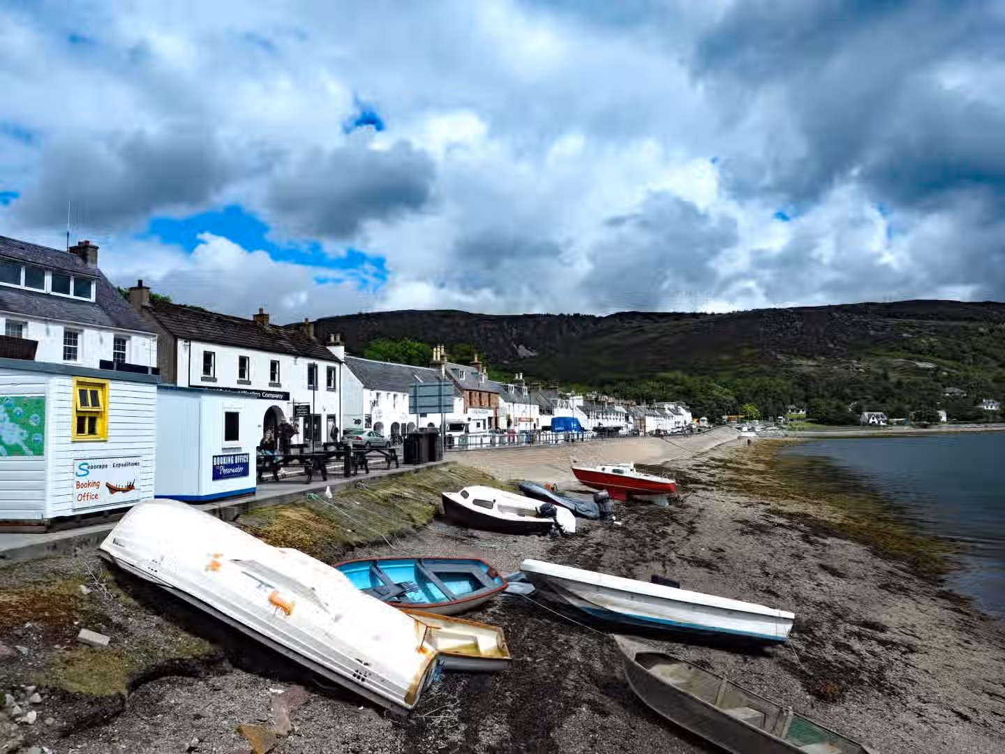 The image depicts a coastal town scene with boats on a beach and buildings in the background. The composition is decent but could be improved. The lighting is adequate, although the cloudy sky creates a somewhat flat light. Sharpness is acceptable but not exceptional. Color accuracy appears reasonable. The image is not particularly photo-realistic, possibly due to the slightly processed look. The boats and buildings are generally in focus. The presence of multiple boats scattered on the beach could be considered slightly cluttered, impacting the overall image quality. The image is not a professionally taken close-up food image, a selfie, a promotional graphic, or an advertisement. The image contains text on the building. Well-composed: 18/25, Well-lit: 12/15, Sharp: 14/20, Color-accurate: 8/10, Photo-realistic: 10/20, Key elements in focus: 3/10.