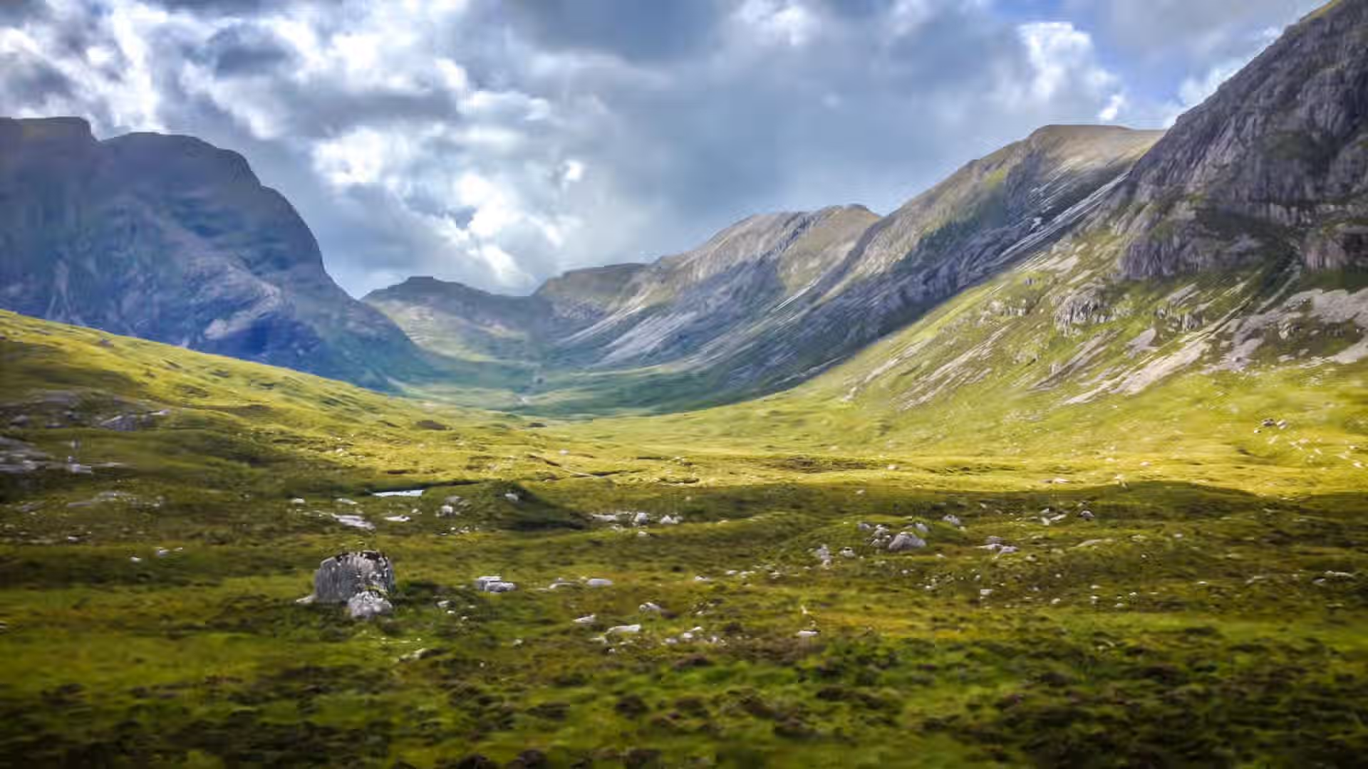 The image showcases a scenic landscape with mountains, a valley, and grassland under a cloudy sky. The composition is decent, capturing the vastness of the landscape. The lighting is somewhat uneven due to the clouds, but generally acceptable. The sharpness is good, although some finer details could be crisper. The colors appear accurate, with the greens of the grassland and the grays of the mountains well-represented. The image is reasonably photo-realistic. Key elements like the mountains and valley are in focus. The image is well-composed, well-lit, sharp, color-accurate, and photo-realistic. However, the overall sharpness could be improved. The composition is good.