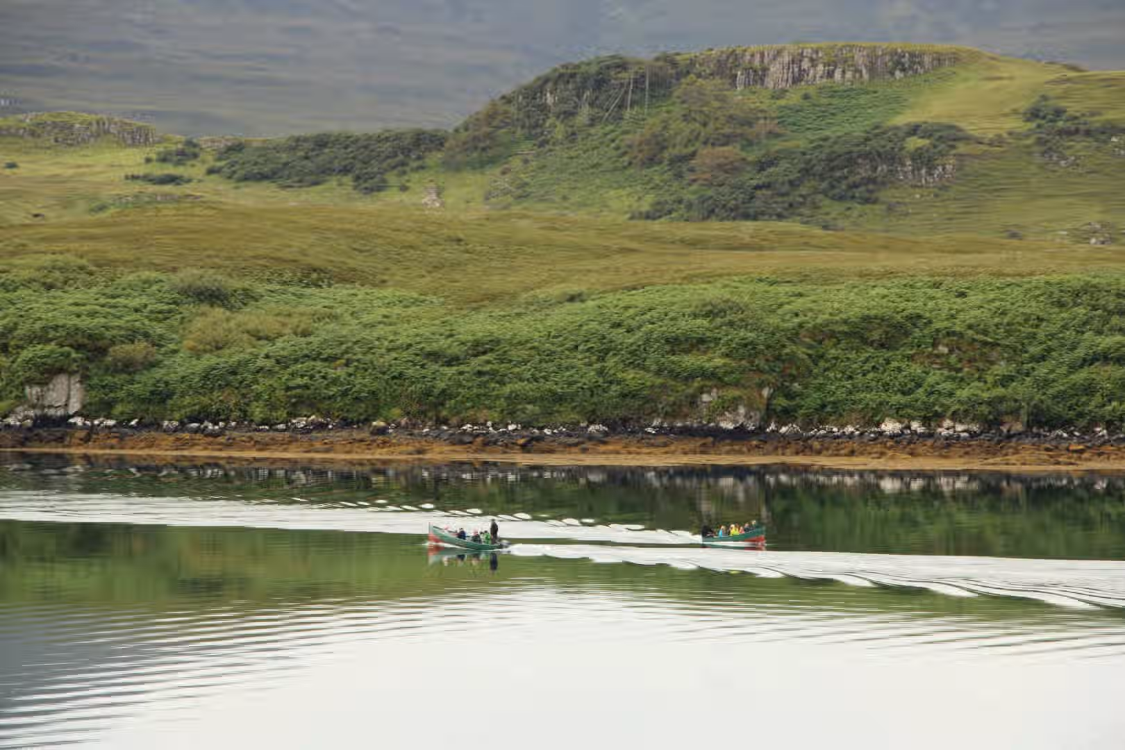 The image shows a landscape with green hills and water. Two boats are visible on the water. The composition is decent, but could be improved by focusing more on one boat. The lighting is soft and even, suggesting an overcast day. The sharpness is acceptable, but not exceptional. The colors appear accurate. The image is reasonably photo-realistic. Key elements, like the boats and the immediate shoreline, are in focus. Overall, it's a pleasant scene, but could benefit from improved composition and sharpness. Well-composed (20/25), Well-lit (13/15), Sharp (15/20), Color-accurate (9/10), Photo-realistic (16/20), Key elements in focus (5/10).