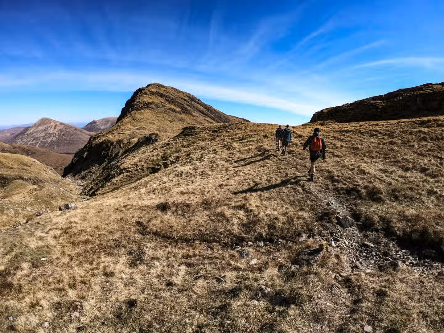 The image depicts a group of people hiking in a mountainous landscape under a clear blue sky. The composition is reasonably well-done, capturing the vastness of the scenery. The lighting is good, with bright sunlight. Sharpness is acceptable, although not exceptionally detailed. Colors appear accurate. The image is fairly photo-realistic. The key elements (mountain and hikers) are mostly in focus. Overall, the image is appealing and conveys a sense of adventure. It could be improved with more dynamic composition and sharper focus.