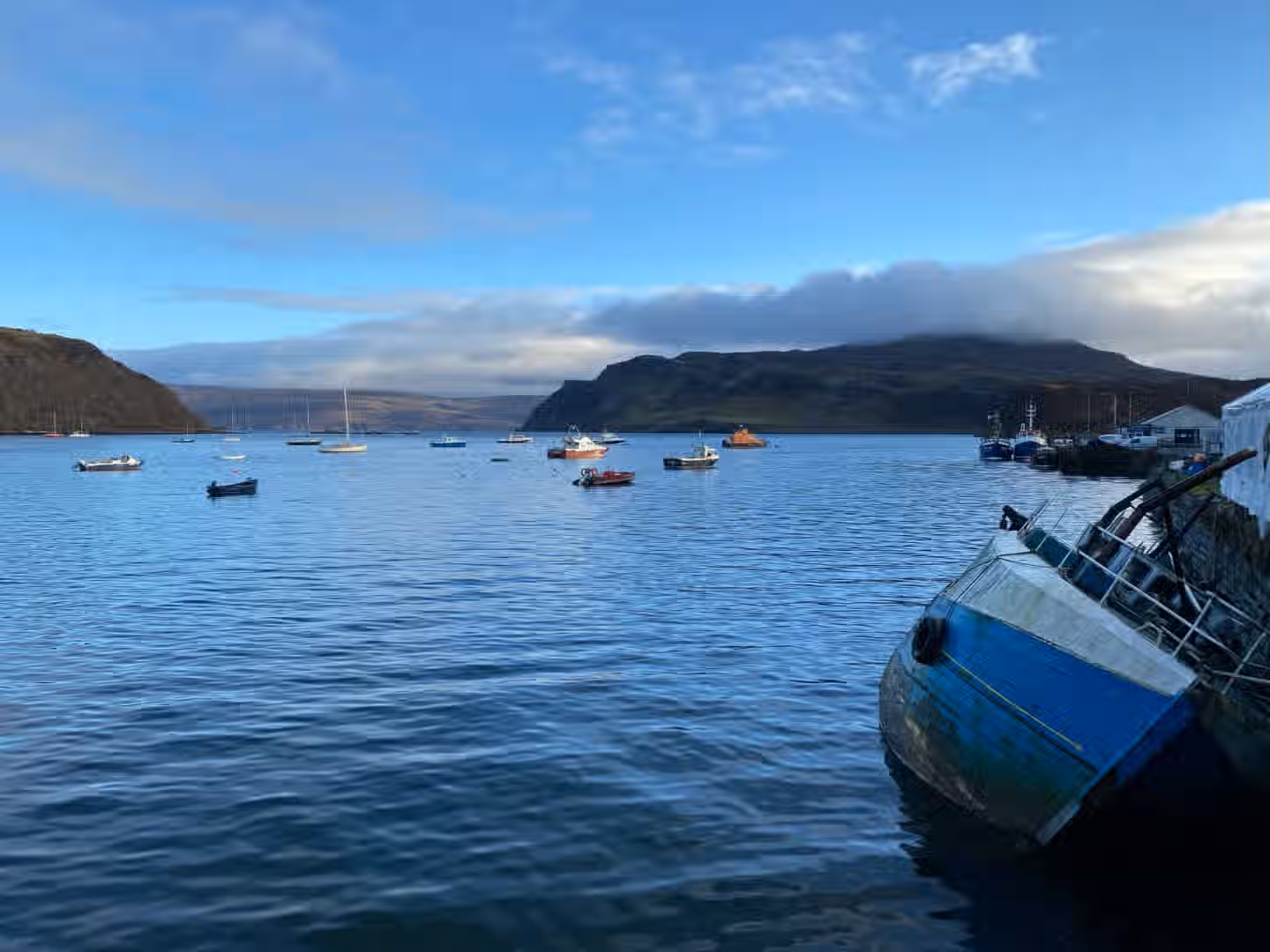 The image depicts a scenic view of boats on the ocean with mountains in the background. The composition is decent, although the boat in the foreground is slightly distracting. The lighting is good, with natural light and color accuracy is acceptable. The image is reasonably sharp, and key elements are mostly in focus. It's a photo-realistic image, but the composition could be improved. The image is not perfect but it is well above average. The boat in the bottom right corner is a bit of a distraction but overall the image is good.