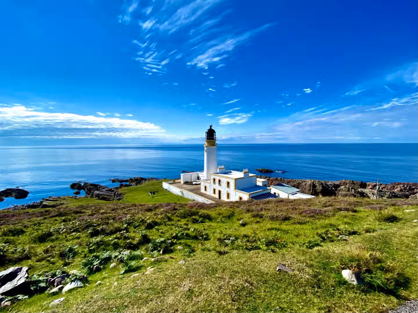 The image of the lighthouse is well-composed, with the subject prominently featured against the backdrop of the ocean and sky. The lighting is bright and clear, enhancing the colors. Sharpness is good, allowing for detail to be seen in the lighthouse structure and surrounding landscape. Color accuracy appears to be well-maintained, with vibrant blues and greens. The image has a photo-realistic quality. The key element, the lighthouse, is in focus. Overall, a high-quality image with minor improvements possible in composition.