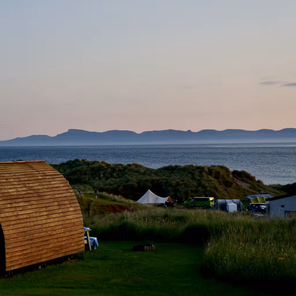 The image depicts a landscape with a cabin in the foreground, an ocean, and mountains in the background. The composition is decent, but the cabin is slightly dark. The lighting is soft, likely taken during dusk or dawn. The sharpness is acceptable, and the colors appear accurate. It has a photo-realistic quality. The key elements, such as the cabin and the distant mountains, are reasonably in focus. The image is well-composed, and the overall scene is appealing. The image could be improved with better lighting on the cabin.
