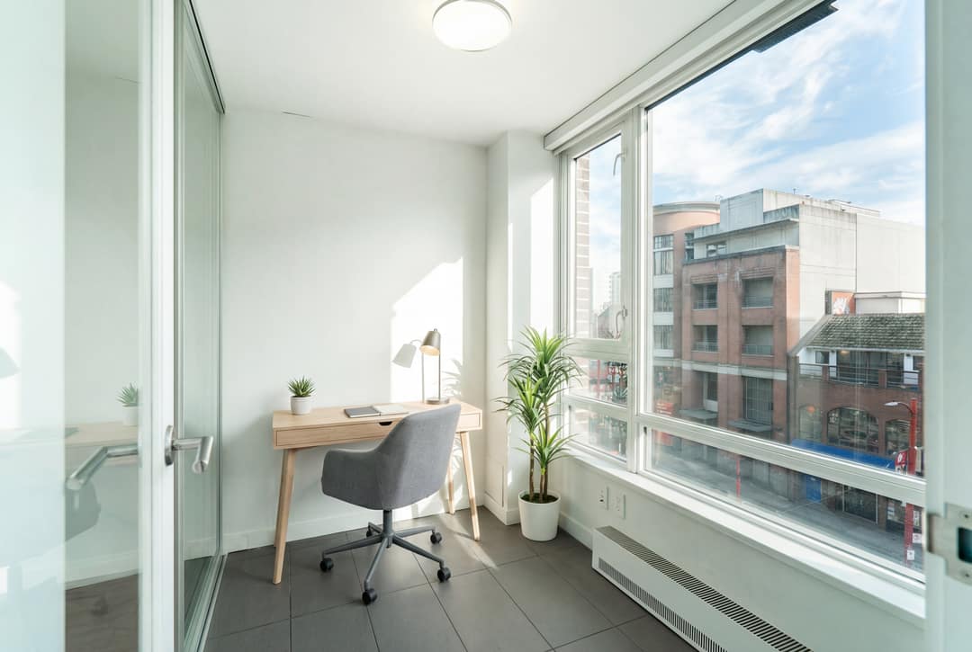 Before and after virtual staging of an empty glass-walled den in Downtown Vancouver, furnished as a bright home office with a desk, chair, and plants overlooking the city streetscape.