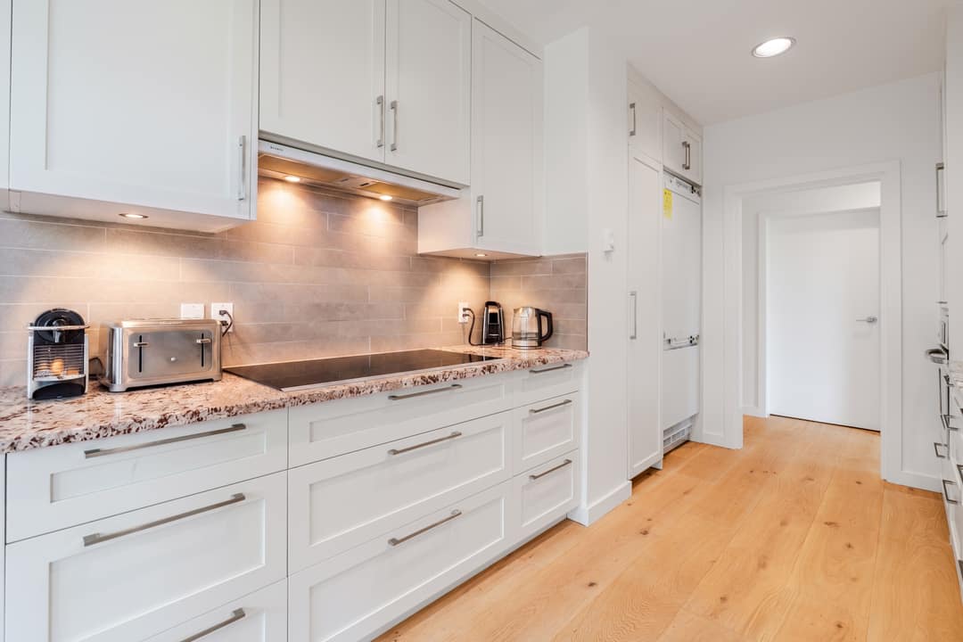Bright kitchen in Vancouver's West End featuring white shaker cabinets, granite countertops, a stone tile backsplash, induction cooktop, under-cabinet lighting, and wide-plank white oak floors.