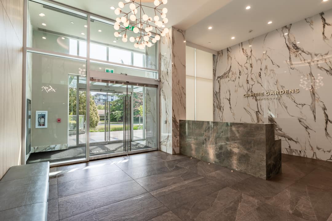 Grand lobby at Cambie Gardens Vancouver featuring a marble feature wall, sculptural globe chandelier, dark stone concierge desk, and automatic glass entry doors.