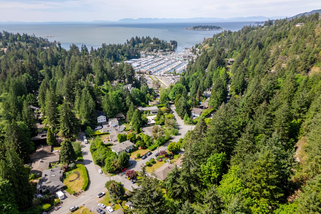 Aerial drone shot above a tree-lined residential street in Horseshoe Bay, West Vancouver, leading to the marina and Howe Sound beyond.