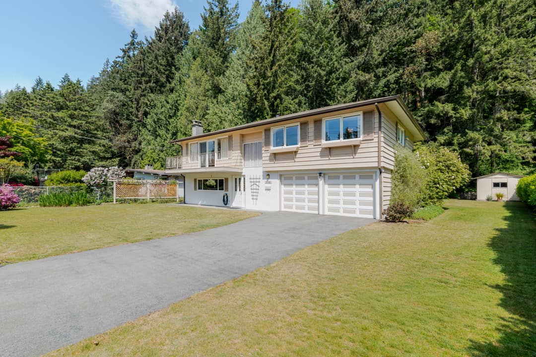 Front exterior of a split-level home in Horseshoe Bay, West Vancouver, showing a double garage, upper balcony, wide driveway, manicured lawn, and a dense west coast forest rising behind the property.