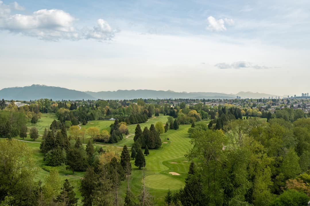 High-floor view from a Cambie Gardens condo overlooking a lush golf course, treetops, and a sweeping mountain range backdrop on a clear spring day.