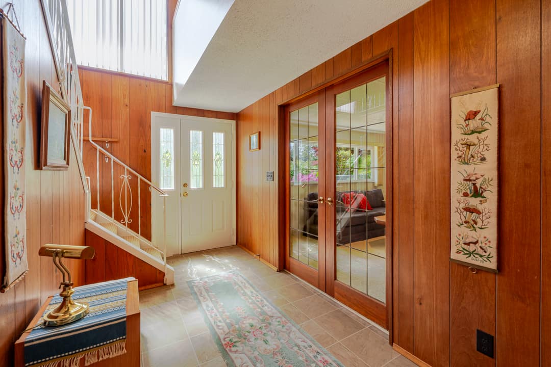 Entry foyer in Horseshoe Bay featuring original cedar wood panelling, a leaded glass front door, leaded glass French doors leading to the living room, and a staircase with wrought iron railing.