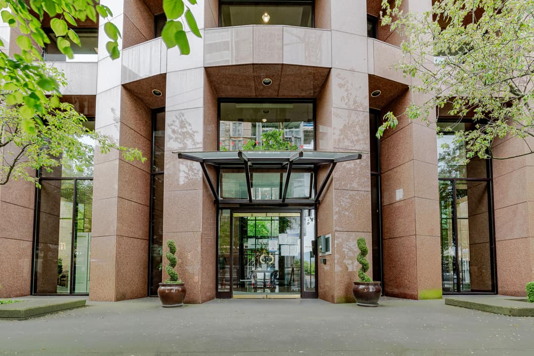 Close-up of a residential tower entrance in Vancouver's West End, featuring a curved rose granite facade, a glass and steel canopy, and symmetrical potted topiary flanking the automatic doors.