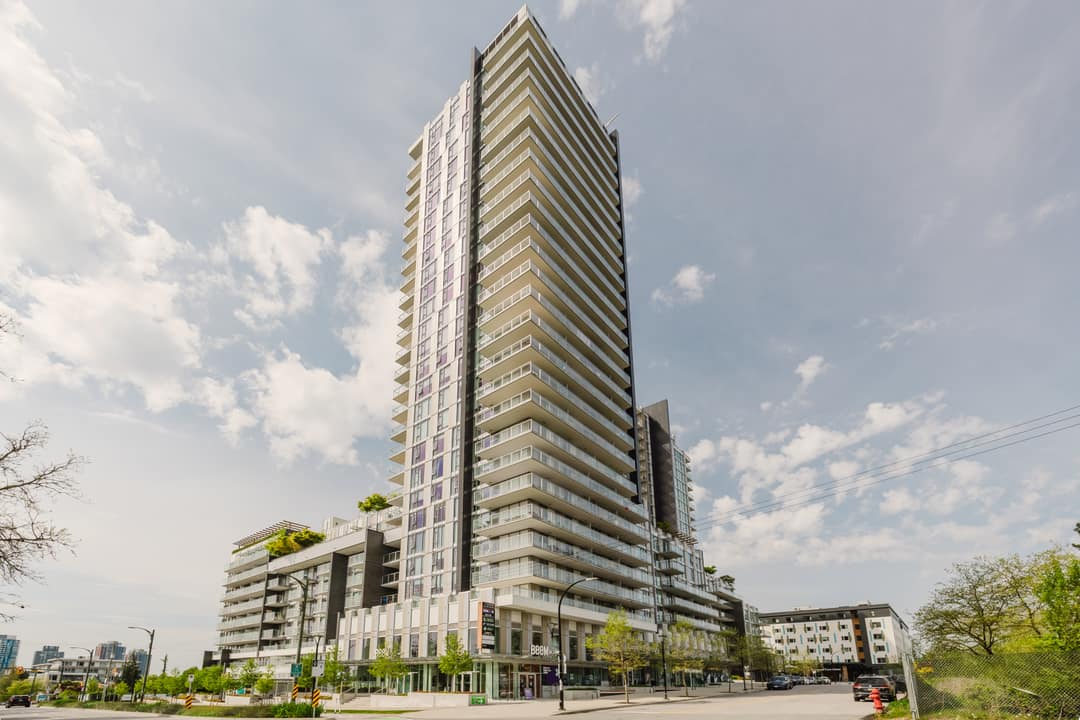 Street-level view of the Cambie Gardens residential tower in Vancouver, showing the full height of the glass and concrete facade against a partly cloudy sky.