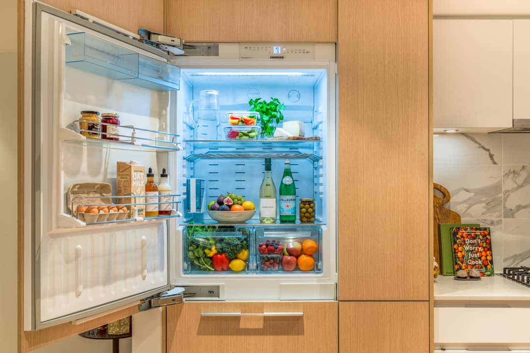 Close-up interior real estate photography of a high-end integrated refrigerator with LED lighting, stocked with fresh groceries, built into custom light-wood kitchen cabinetry.