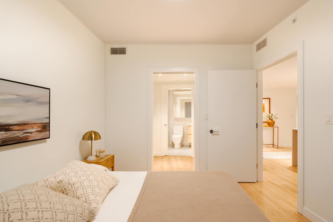 Interior real estate photography of a bright master bedroom in North Vancouver, looking through an open door into a modern white ensuite bathroom with large-scale tiling.