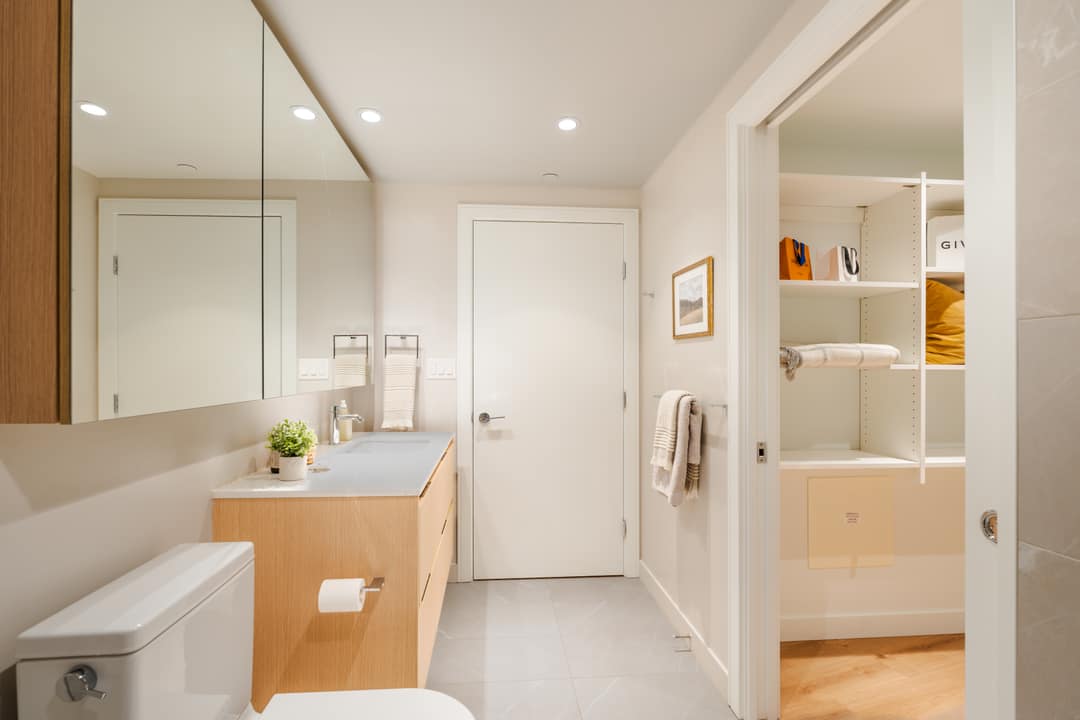 Interior real estate photography of a modern ensuite bathroom in North Vancouver, showing a light-wood vanity and an open door to a custom walk-in closet.