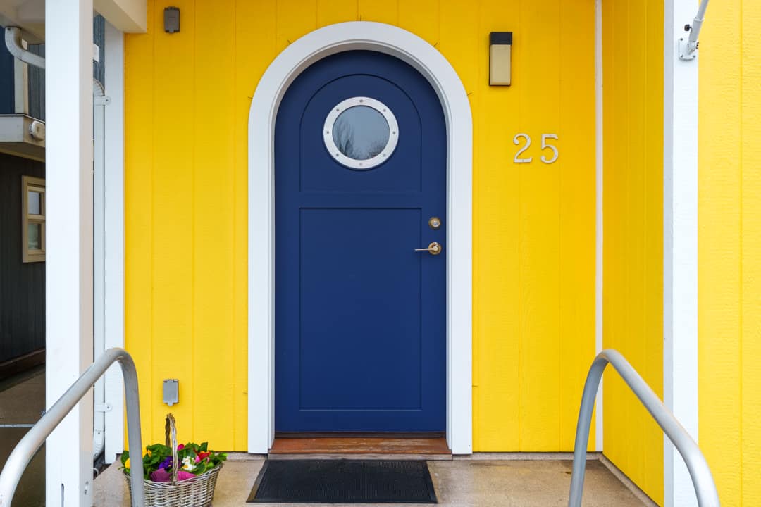 Exterior real estate photography of a luxury floating home in North Vancouver, featuring a vibrant yellow facade and a navy blue front door with a circular porthole window.