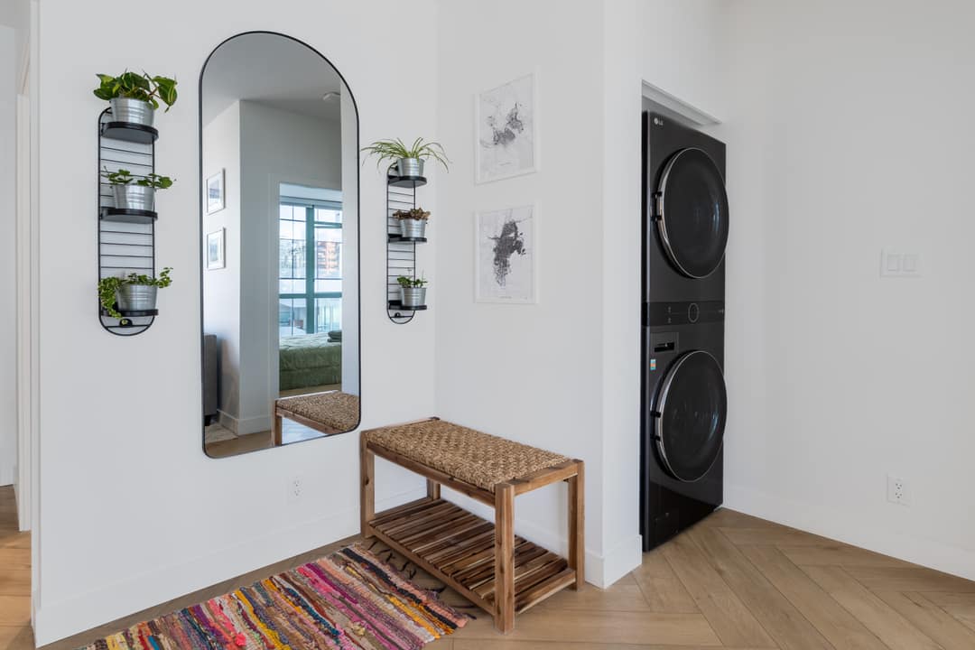 Interior real estate photography of a compact hallway in a Downtown Vancouver Airbnb, featuring a large arched mirror, a wooden bench, and integrated black-and-white stacked laundry units.