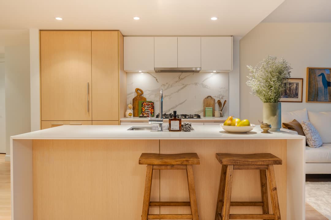 Bright interior real estate photography of a modern kitchen in North Vancouver featuring light oak wood cabinetry, marble slab backsplash, and a clean white island with wooden bar stools.