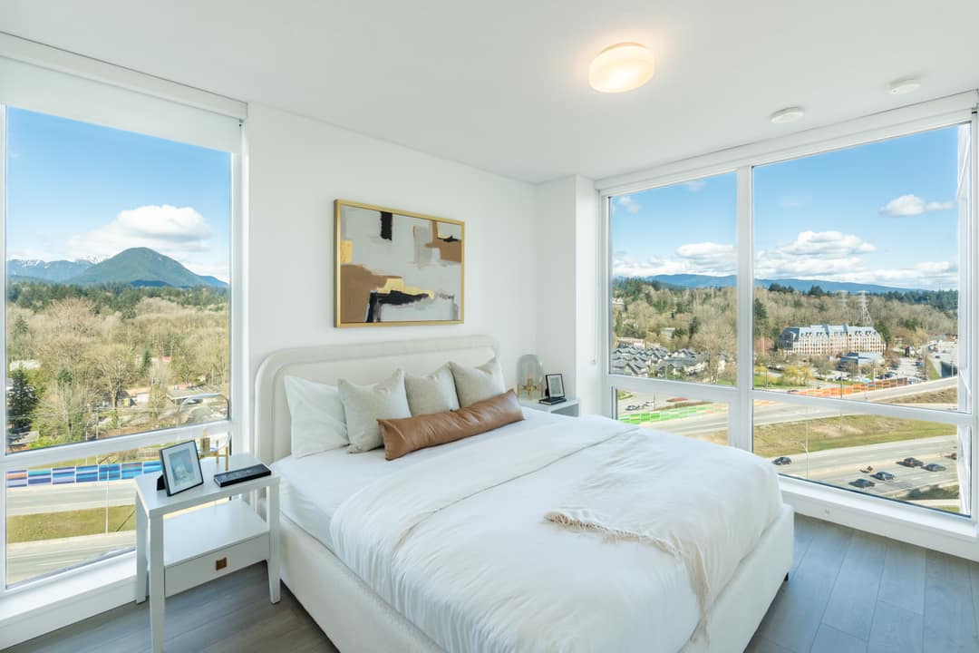 Interior real estate photography of a sun-drenched primary bedroom in North Vancouver, featuring expansive floor-to-ceiling windows with elevated community views, clean white bedding, and modern minimalist decor.