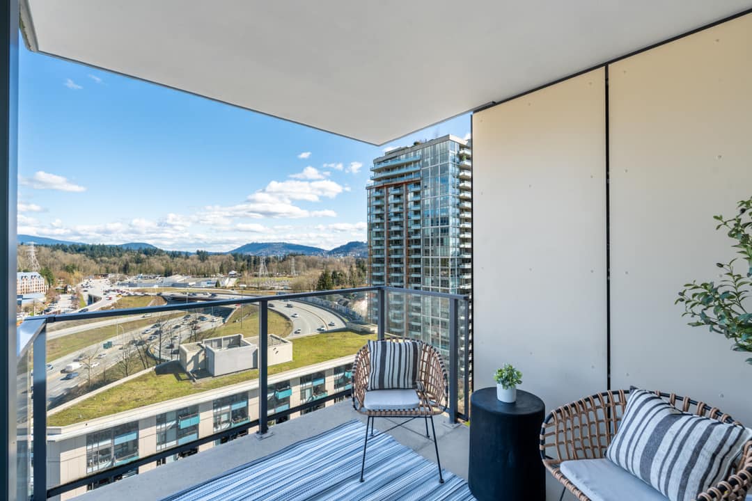 Interior real estate photography of a sun-drenched primary bedroom in North Vancouver, featuring expansive floor-to-ceiling windows with elevated community views, clean white bedding, and modern minimalist decor.