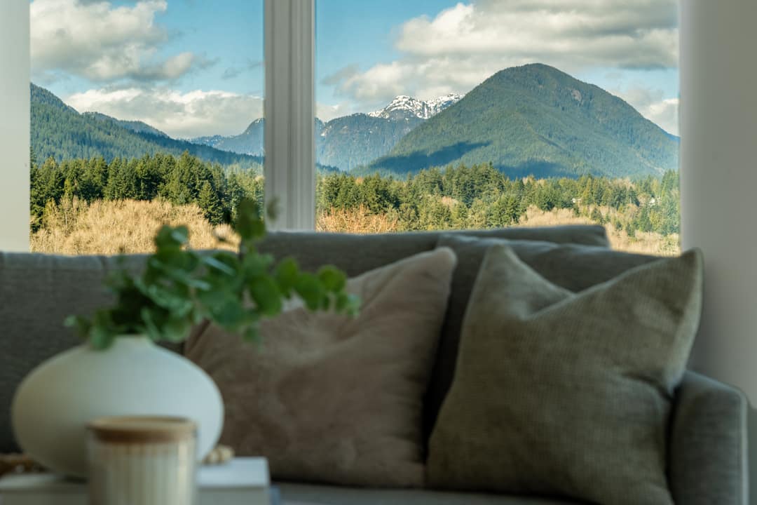 Interior real estate photography of a modern living room in North Vancouver, featuring floor-to-ceiling windows that provide a breathtaking view of the dense green forest and snow-capped mountain peaks.