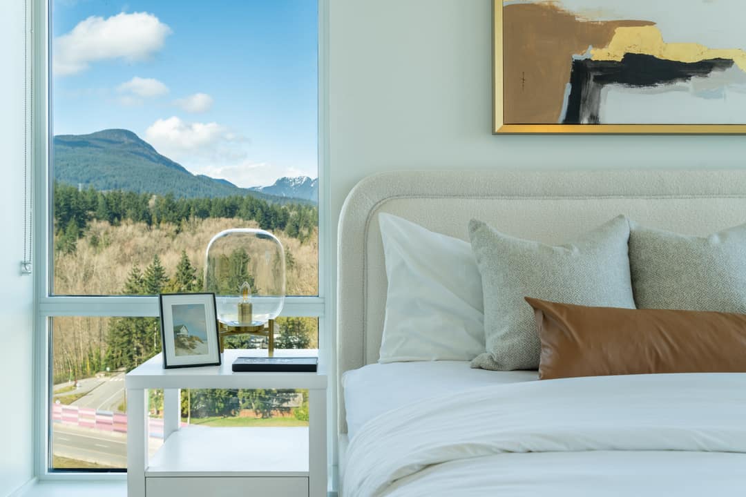 Interior real estate photography of a bright primary bedroom in North Vancouver, featuring a modern bed with neutral tones, a minimalist nightstand, and a large window offering a stunning view of the North Shore mountains and forest.