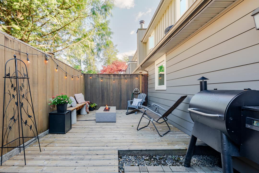 Backyard deck in Squamish styled with a concrete fire pit, lounge chairs, string lights, potted flowers, and a Traeger smoker, captured in warm evening light.