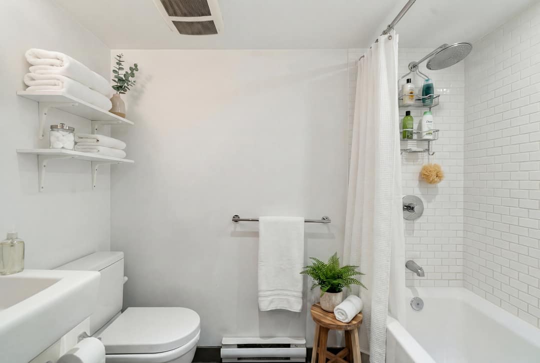 Before and after virtual staging of a plain white bathroom in Downtown Vancouver, dressed with folded towels, a fern, wood stool, and shelf decor to create a clean, spa-like presentation.